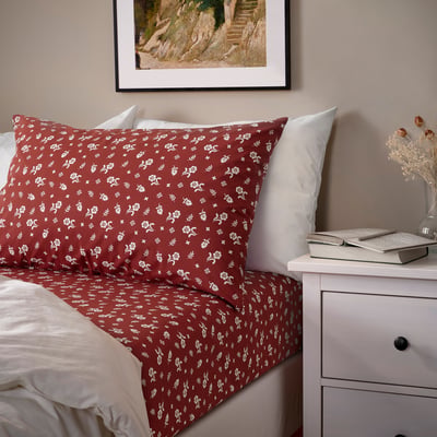 A maroon TÅGSÄCKMAL bed sheet with white florals, beside a drawer with a laptop and dried flowers, framed picture above.