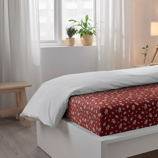 Bed with white comforter and red patterned sheet near window with plants.