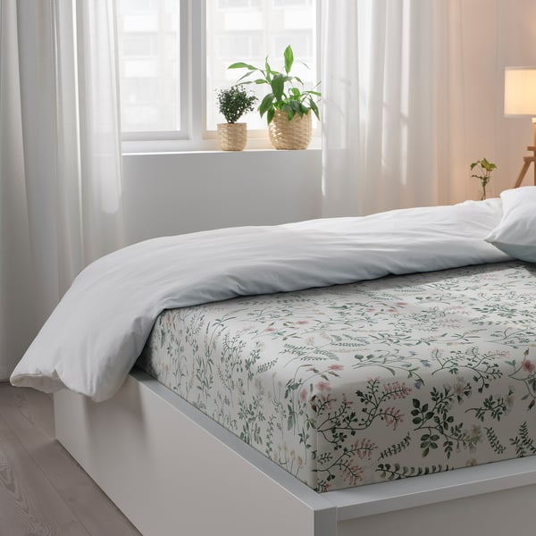 A cosy bed with white sheets and floral print, two potted plants on the windowsill, and soft natural light.