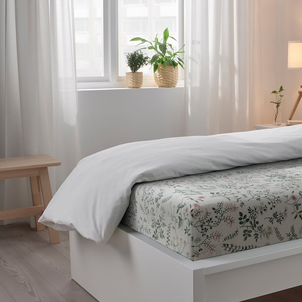 Cosy bedroom with white bed, floral sheet set, two potted plants on windowsill, wooden floor, and bench.