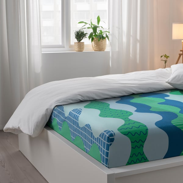 Bright bedroom with white bed, blue-green wave sheets, two plants on windowsill.