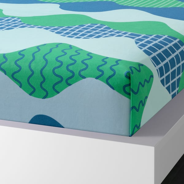 Bed sheet set with blue-green waves. Soft 100% cotton, machine-washable. Envelope pillowcases shown on white bed.