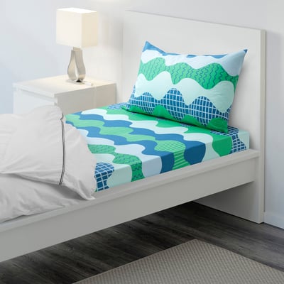 Blue-green wave bedding set on white bed, 100% cotton. Small lamp on bedside table, plain wall.