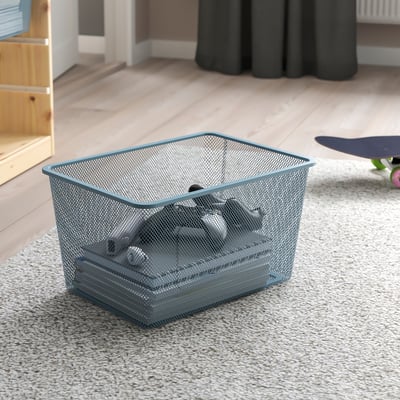 Mesh storage bin on carpeted floor, containing several items, designed for easy access and organisation. Ideal for indoor use with TROFAST frames.