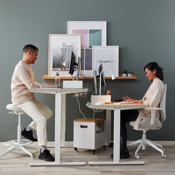 Two people at adjustable desks with computers and crank handles. Artwork on wall behind.