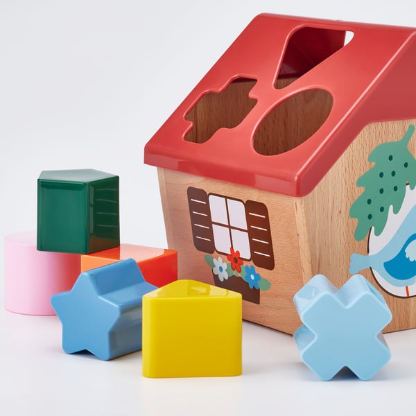Brightly coloured wooden blocks in various shapes sit near a wooden birdhouse-shaped toy with cut-outs, teaching shapes and colours.
