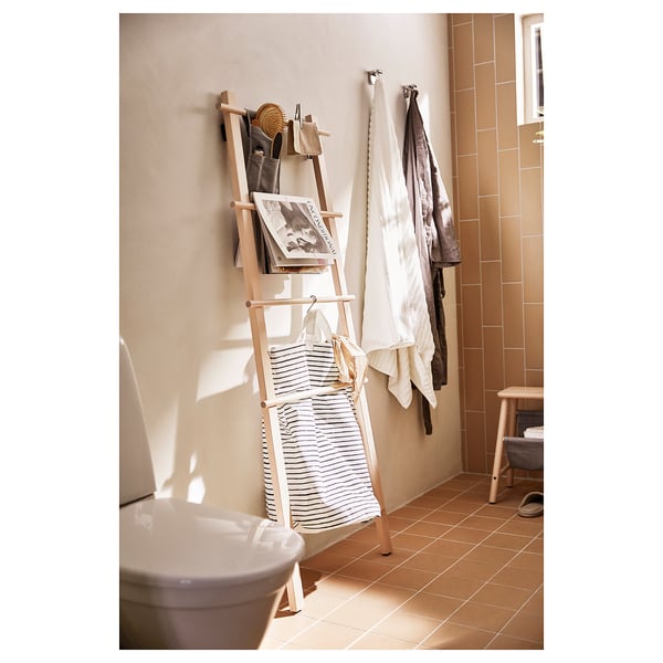 A VILTO birch wooden towel stand, natural in colour, with adjustable feet, holding striped towels and a magazine in a bathroom setting.