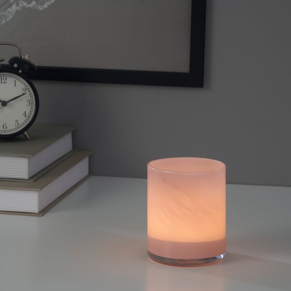 A lit candle in a pink glass holder sits on a white surface, accompanied by a stack of books and an old-fashioned clock.