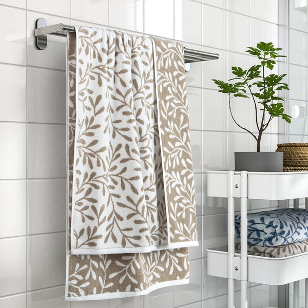 White towel with brown leaf pattern hangs on silver rail, next to green plant on white stand.