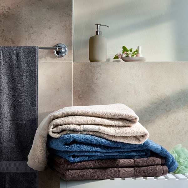 A neat stack of folded towels in beige, blue, and brown on a bathroom counter, accompanied by a soap dispenser and a small pot of flowers.