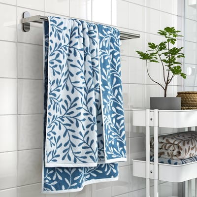 Blue towel with leaf pattern hangs on rail near white shelf with folded towels and plant.