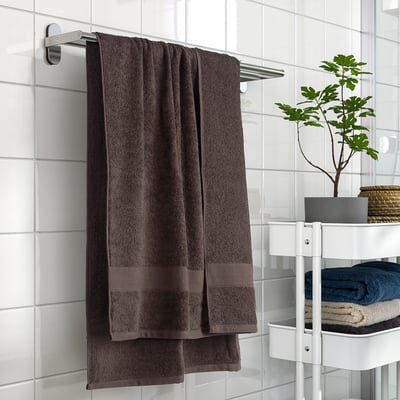 Brown VITBACKEN bath towel hanging on a rack, 100% cotton, medium-thick & soft. Nearby, folded towels & a potted plant on white shelf.