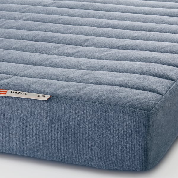 Close-up of a firm mattress with textured blue fabric. Tags show the brand VOGNILL and mention eco-friendly materials.