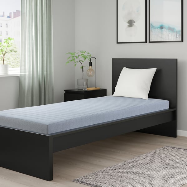 Black mattress on bed, natural materials, beside a lamp and plant.