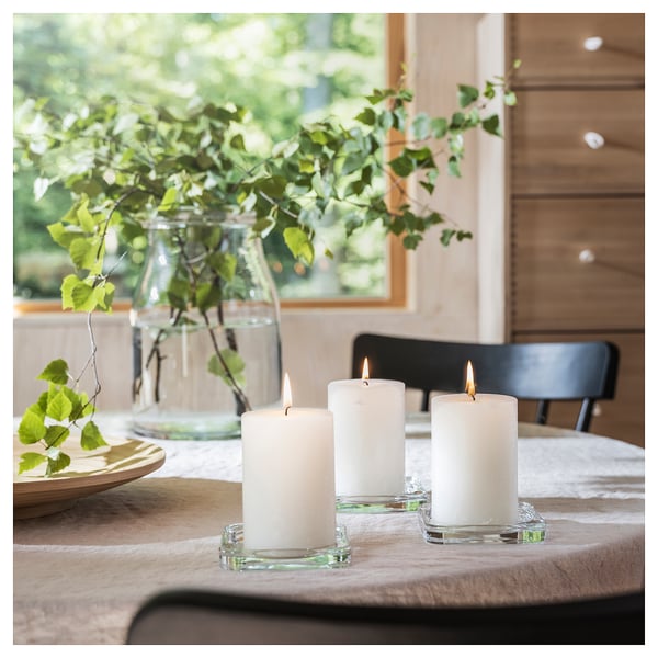 Calm table setting with three white ADLAD candles in glass holders, a vase of green leaves, wooden plate, on fabric with natural light.