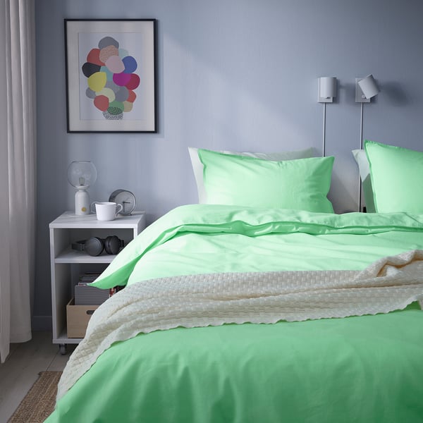 Bright bedroom with light green ÄNGSLILJA cotton duvet set, white accents, and colourful wall art; bed is covered with green duvet and pillowcases.