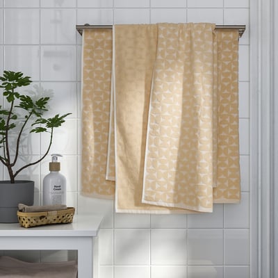 Beige towels with white pattern hang on silver rack; green plant and hand cream on counter, folded towels below.