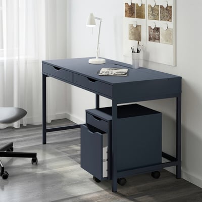 Grey ALEX desk with drawers, lamp, notebook, pen holder in minimalist room. On wheels, next to black chair, wall with abstract art.