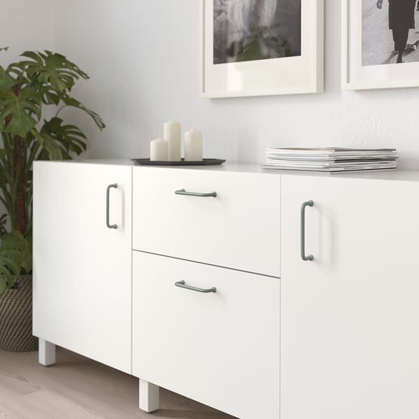 White BAGGANÄS sideboard, minimalist design, 2 drawers, 2 doors with silver handles. Between pictures and plant.