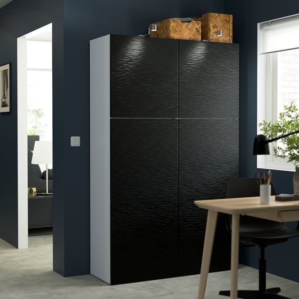 Modern, tall black BESTÅ storage cabinet with woven baskets on top, next to a wooden table and chair in a contemporary room.