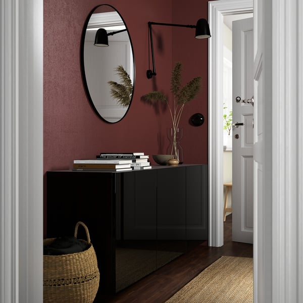 Modern entryway with BESTÅ cabinet, mirror, books, plant. Burgundy wall, wicker basket, and rug create cosy, stylish space.