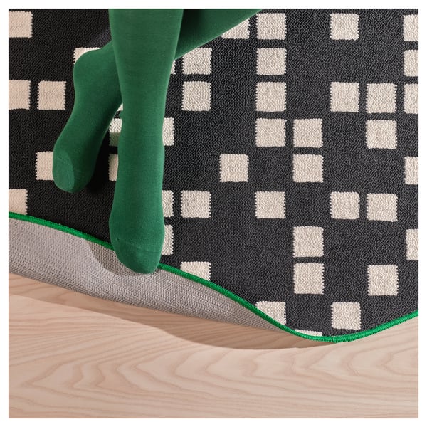 A persons feet wearing green socks on a gaming rug with a black and white pixel pattern, emphasising the rugs quality and design.