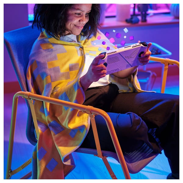 A person sits on a colourful chair, wrapped in a cosy pixel-patterned BRÄNNBOLL throw blanket, holding a gaming console.