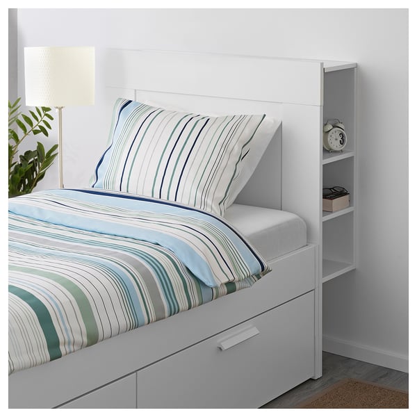 A white BRIMNES headboard with built-in shelves and storage compartments for books and items, attached to a bed with a striped comforter and pillows.