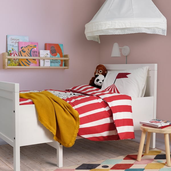 Kids white bed with red-striped circus-themed duvet, yellow blanket, stuffed animal, bookshelf with books, small stool, colourful rug, pink walls.