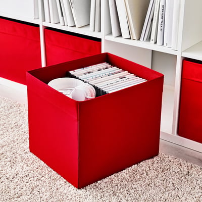 Red fabric storage box, filled with items, placed on textured beige carpet. Part of kallax shelving unit with white books.