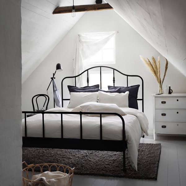Black DVALA bed, white linens, table lamp, chair, dresser with grasses, rug, basket, slanted beams.