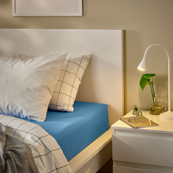 Bed with blue DVALA cotton fitted sheet, white pillows, bedside table, flower, lamp, on beige wall.