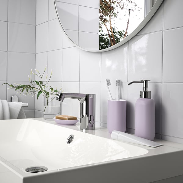 Modern bathroom with EKOLN light purple soap dispenser and toothbrush holder on counter, mirror reflecting tree, and white sink.
