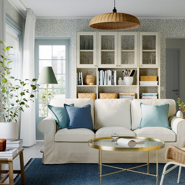 Bright living room with light-colored EKTORP sofa, blue pillows, glass coffee table, bookshelf, and large window.