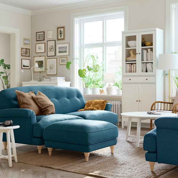 Modern living room with blue ESSEBODA sectional sofa, gold pillows, white cabinet, rug, and large window with plants.