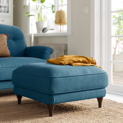 Bright blue ESSEBODA footstool with wooden legs, folded mustard blanket on top, cosy living room setting.