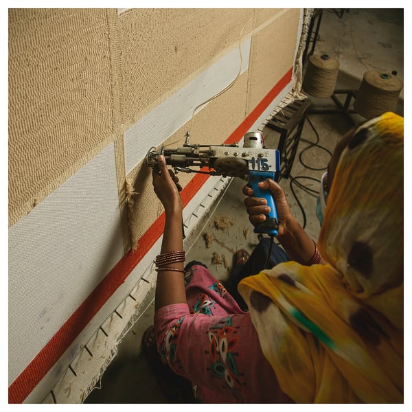 A person is sewing a beige and red striped rug by hand. The person uses a blue tool and wears colourful clothing.