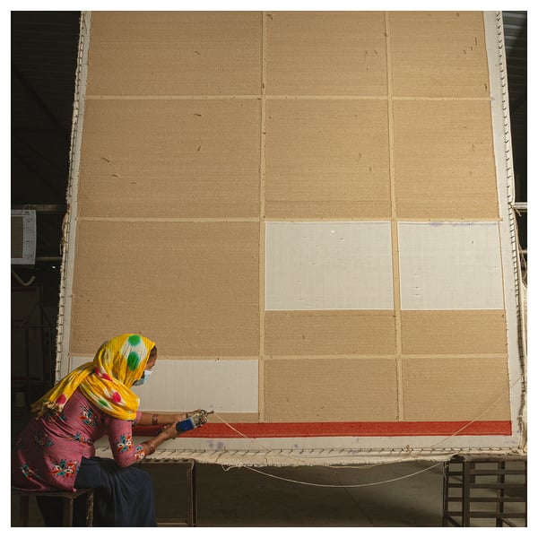 A person weaves a beige and red rug, wearing a colourful headscarf.