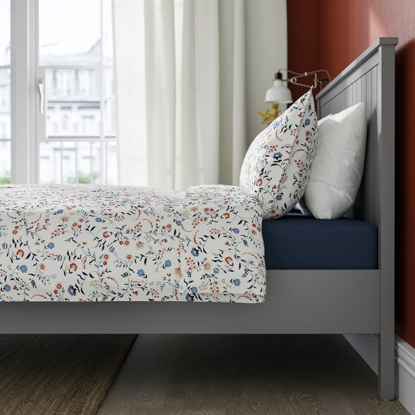 A grey metal bed frame with a floral-patterned bedspread and a white pillow. The frame has airy legs and an adjustable slat support for mattresses.