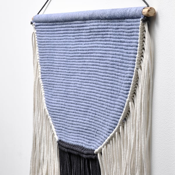 Hanging wall textile, blue-gray with white fringe, natural wooden top, neat and textured, handmade quality evident.