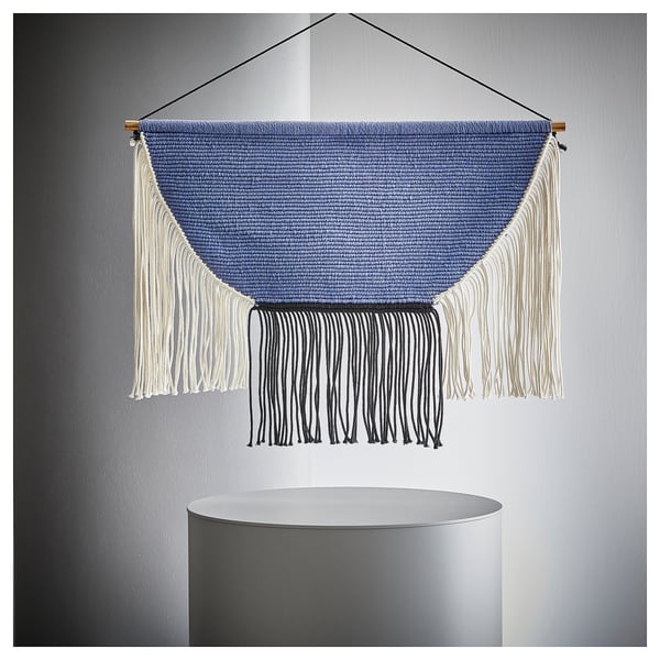 A HÄNGALM wall hanging in blue and white. Its triangular with fringes at the bottom, suspended by a wooden ring.