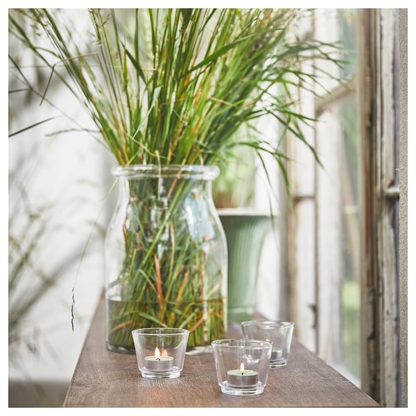 Three lit candles in clear glass holders beside a large glass jar with green grasses on wooden shelf near window.