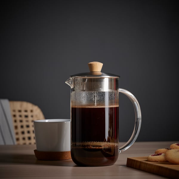 Clear glass cafetière with wooden lid and handle sits on a table next to a white mug and a plate of cookies.
