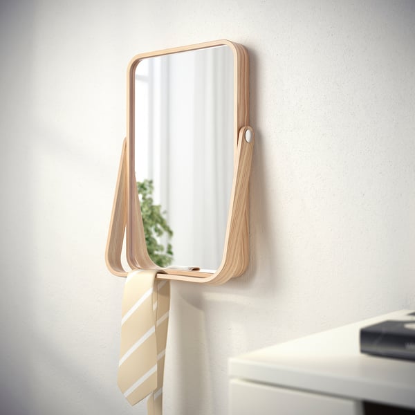 Wall-mounted mirror with hanging shelf for accessories. Light wood frame, reflecting room.