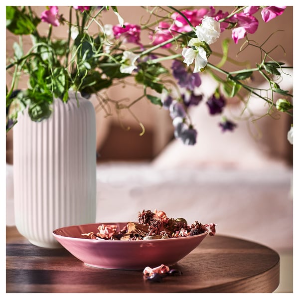 A red bowl of fragrant potpourri next to a white, ribbed vase with pink and purple flowers on a wooden surface.