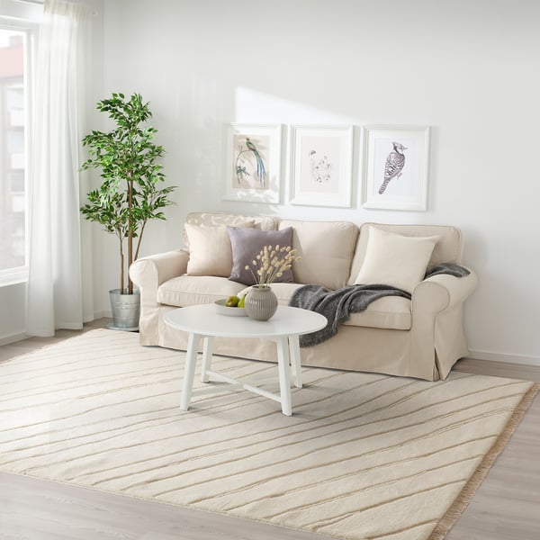 A living room with a JULGRANSGRÄS beige rug, a beige sofa, and nature-themed wall art.