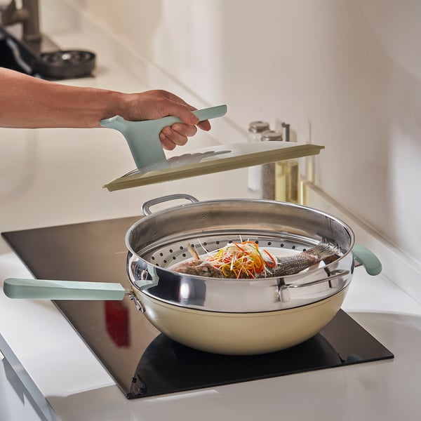 A hand places a green KLOCKREN steamer insert into a shiny pot on a stove, demonstrating its function to steam food.