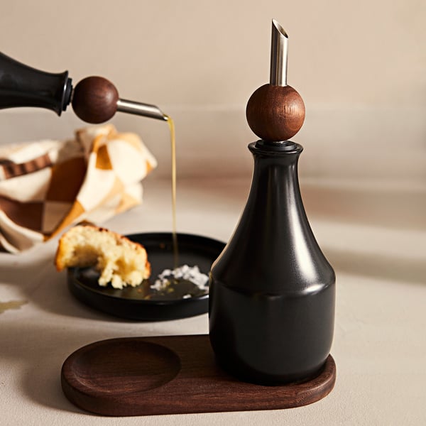 Black KRÖSAMOS oil dispenser with wooden stop on tray. Food in bowl beside.