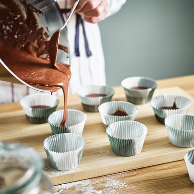 Person pouring batter into cupcake liners on wooden board.