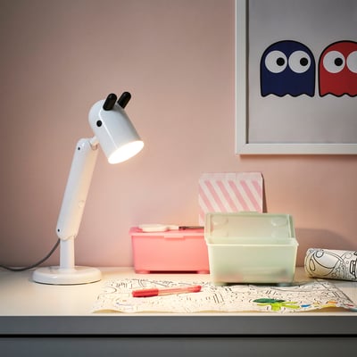 KRUX lamp, white, adjustable. Small, rounded body, flexible arm. On a desk with crafts.